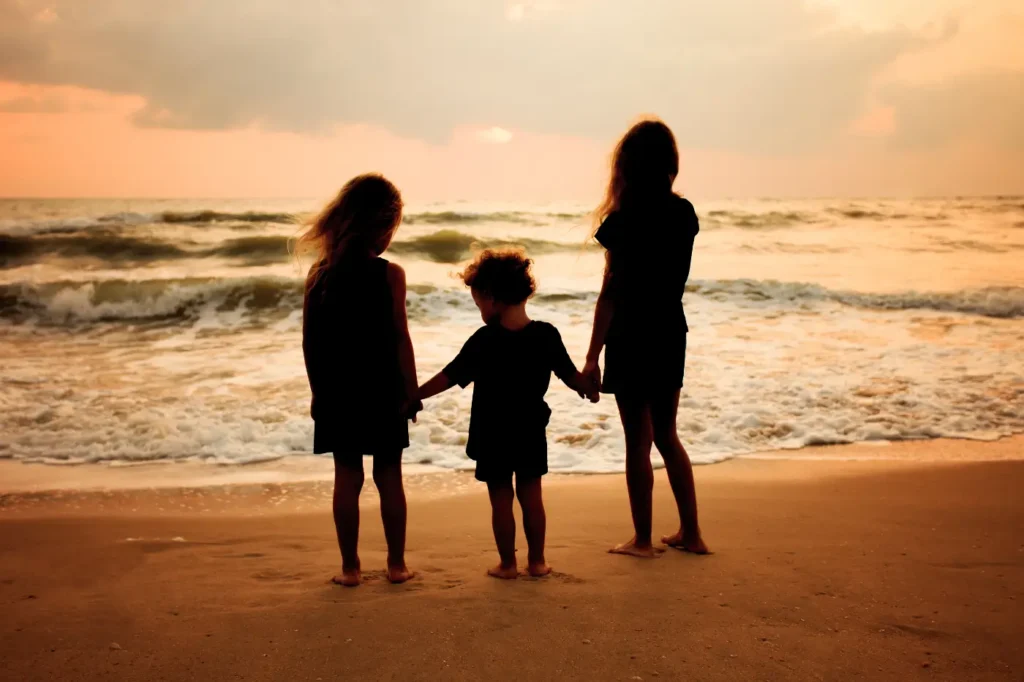 Three children hold hands on sandy beach looking out to sea